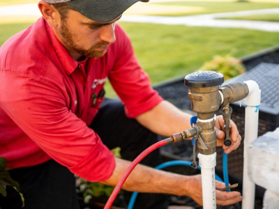 A large, bright red double check valve backflow preventer assembly installed outdoors on a concrete pad, surrounded by green shrubs and mulch.