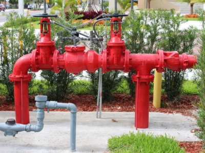 A large, bright red double check valve backflow preventer assembly installed outdoors on a concrete pad, surrounded by green shrubs and mulch.