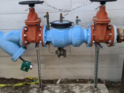 A blue and orange Reduced Pressure Zone (RPZ) backflow preventer assembly mounted on metal struts against a light-colored stone wall.