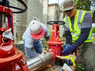 Two technicians in safety gear and hats work on a large red and stainless steel backflow prevention assembly installed outdoors near a residential building.