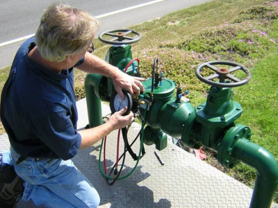 A technician in a blue shirt and jeans is kneeling on a metal platform, performing a test on a large green backflow prevention assembly using a specialized pressure gauge.