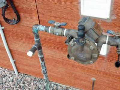 A residential brass backflow prevention assembly with two blue-handled ball valves and black rubber test cock covers, installed outdoors against a wood-paneled wall.