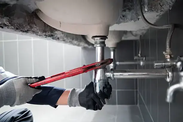 Professional plumber using a large red pipe wrench to perform garbage disposal repair in Fort Lauderdale, working on the undersink piping.
