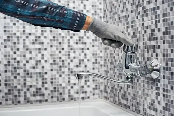 A plumber's gloved hand adjusts a chrome faucet with running water against a gray mosaic tile wall.