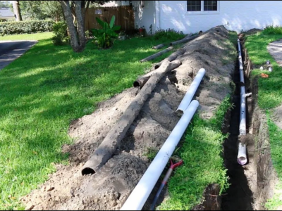 A long, excavated trench for a sewer line repair in Fort Lauderdale featuring new white PVC piping being installed alongside old, corroded cast iron pipes.