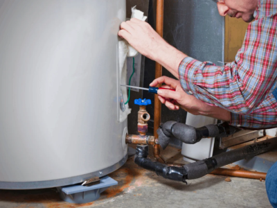 A technician in a red plaid shirt uses a screwdriver to adjust the thermostat or wiring of a residential electric water heater while performing maintenance.