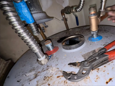 A top-down, close-up view of a corroded silver water heater tank. A pair of red-handled channel lock pliers rests on the tank next to various plumbing connections, including flexible corrugated metal hoses and a brass T-shaped temperature and pressure (T&P) relief valve.