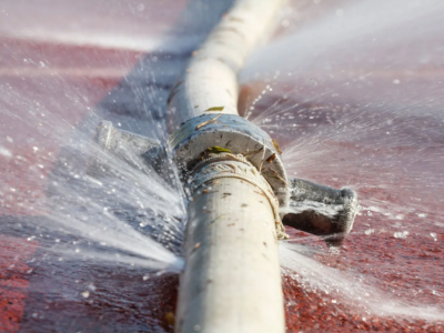 Close-up of a burst water main pipe spraying water under high pressure, requiring a fast-response emergency plumber Fort Lauderdale.
